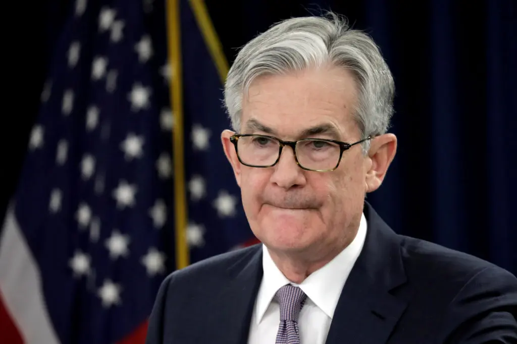 Jerome Powell speaking at a press conference with U.S. flag in the background.