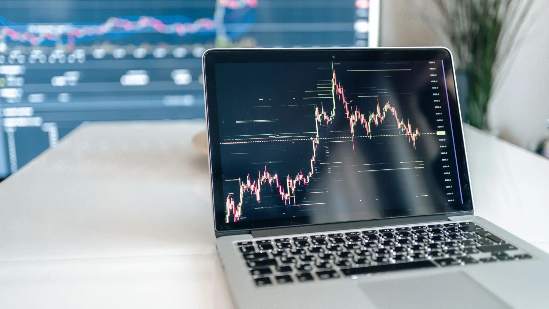 A laptop displaying a financial candlestick chart on a dark screen with another monitor in the background