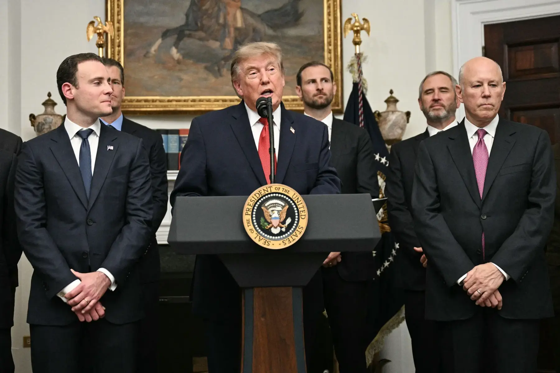 Donald Trump speaking at a podium with the Presidential Seal, surrounded by men in suits