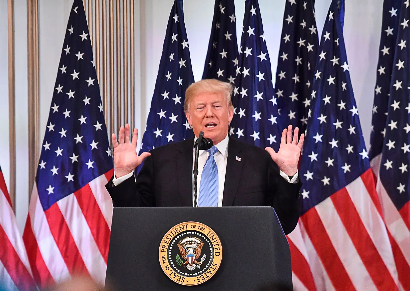 Donald Trump speaking at a podium with the Seal of the President of the United States and several US flags.