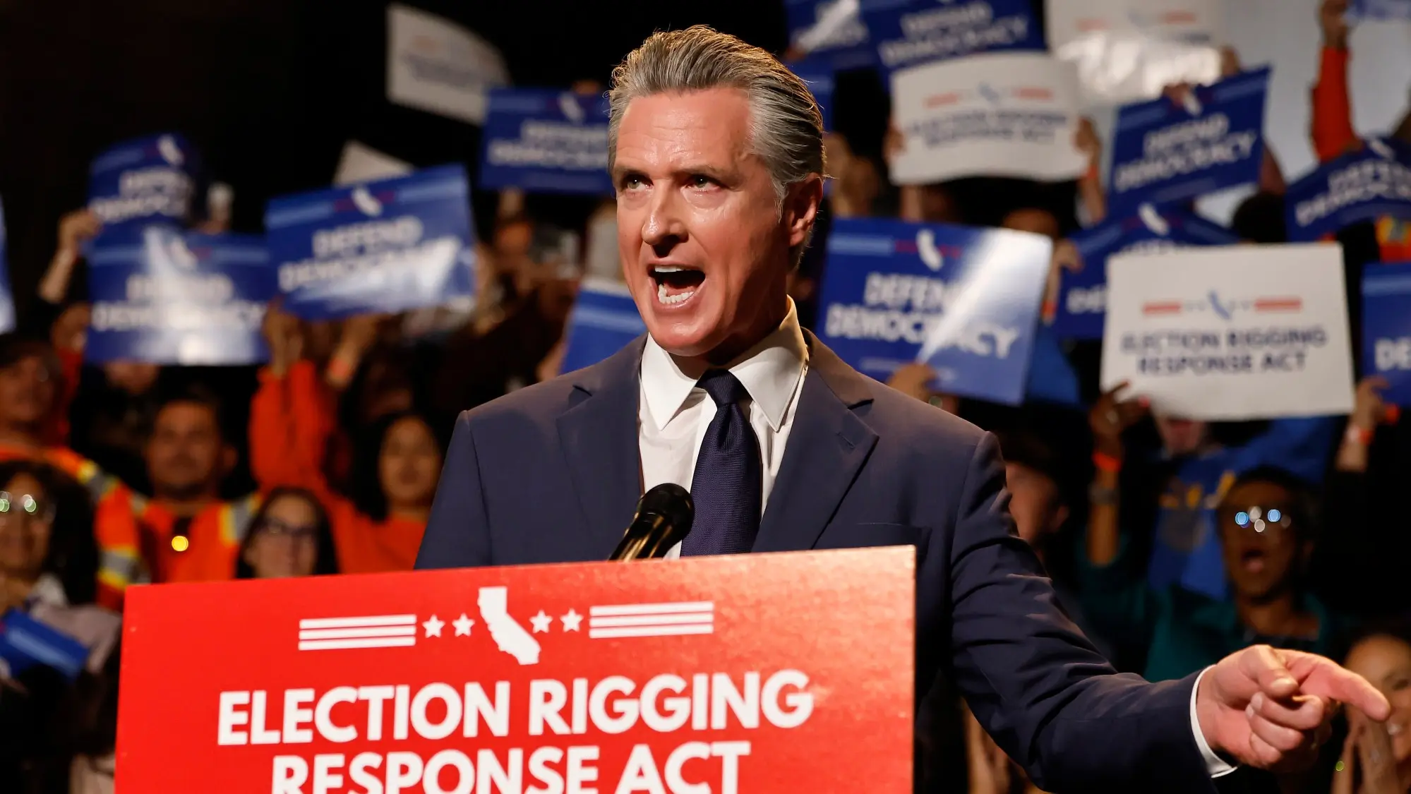 Governor Gavin Newsom speaking at a podium with a red sign reading Election Rigging Response Act