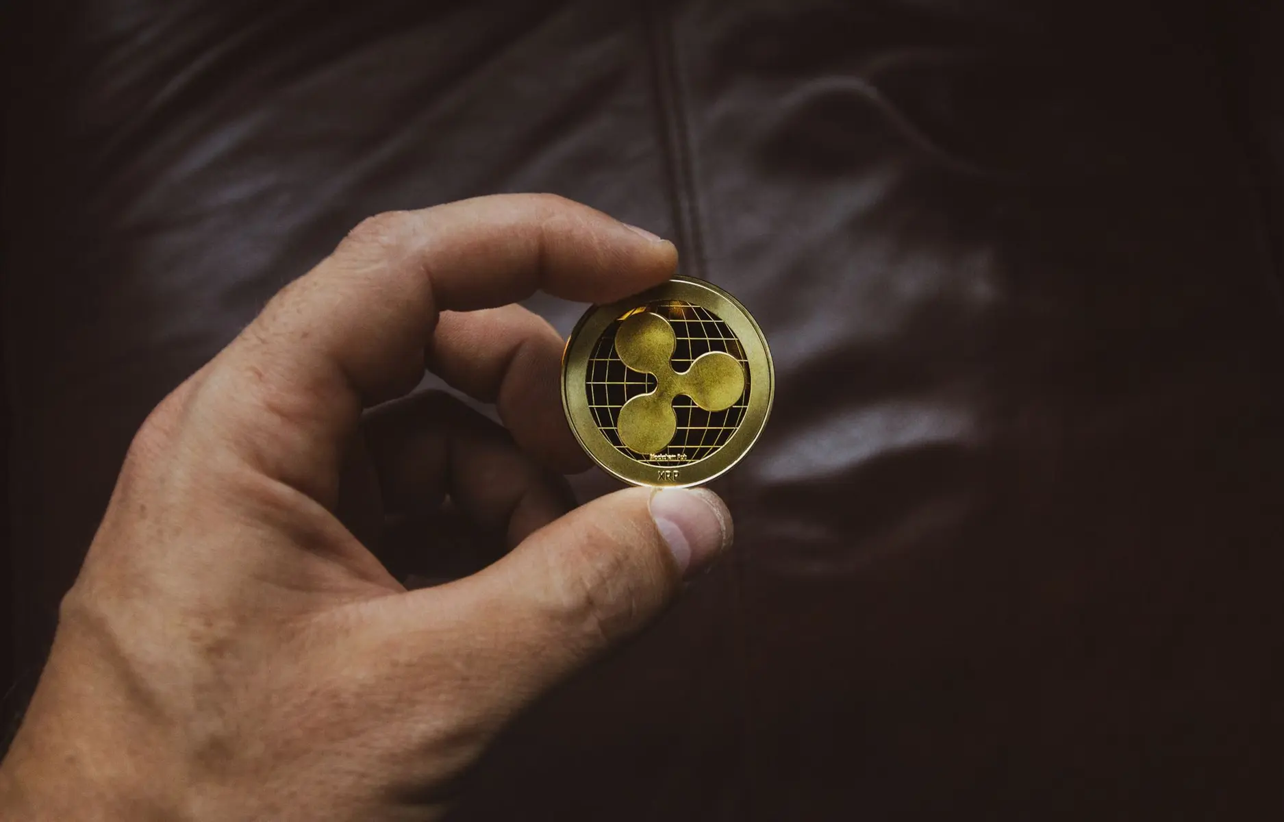 A hand holding a physical gold-colored coin featuring the XRP cryptocurrency logo.