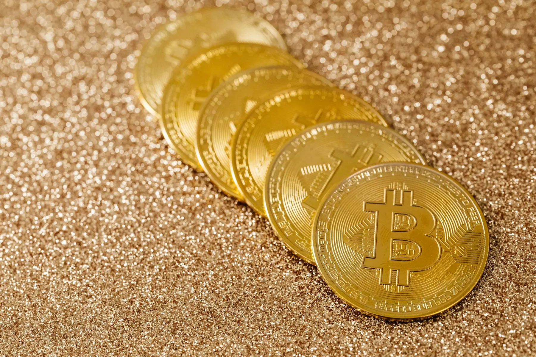 A row of gold-colored Bitcoin physical coins resting on a shimmering gold glitter surface.